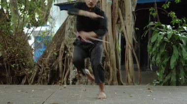 Geleneksel kıyafetli bir dövüş sanatçısı, ağaçlar ve yeşilliklerle çevrili doğal bir ortamda bir duruş sergiler. Sahne, kültürel dövüş sanatlarının özünü yansıtıyor..