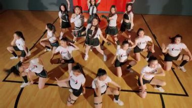 Bir grup genç dansçı bir spor salonunda koreografi gösterisi yapıyor. Aynı kıyafetleri giyiyorlar ve çeşitli dans pozları veriyorlar. Zemin ahşap ve arka planda bir basketbol sahası var..