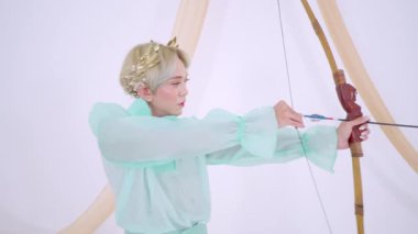 Çiçekli taçlı, nane yeşili kıyafetli biri yaylı bir oka nişan alıyor. Arka plan yumuşak ve nötr. Okçunun duruşuna ve kıyafetine odaklanır..