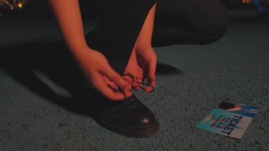 Mavi bir yüzeye diz çöküp siyah bağcıklarını bağlayan bir insan. Yakınlarda, yerde açık bir dergi var. Işıkların ve yeşilliklerin bulanık arkaplanı..