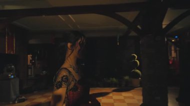 Bir çift, renkli elbiseli ve takım elbiseli bir kişiyle şenlikli bir etkinlikte dans ediyor. Ayar yumuşak ışıklarla aydınlatılıyor, romantik bir atmosfer yaratıyor..