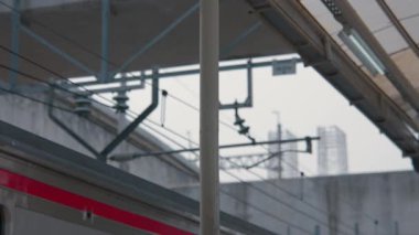 Arka planda bulanık tren rayları ve üstten yapıları olan bir tren istasyonu direğinin yakın çekimi. Sahne kentsel ve endüstriyel, modern ulaşım altyapısı sergileniyor.