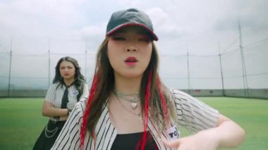 Arka planda bulutlu bir gökyüzü olan yeşil bir alanda dans eden ve alkışlayan iki genç kadının olduğu canlı bir açık hava sahnesi. Bir kadın kep takıyor ve uzun saçlı ve kırmızı röfleli..