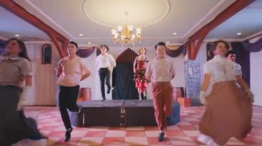 Bir grup dansçı zarif dekorlu geniş bir salonda sahne alıyor. Dansçılar çeşitli kıyafetler giyip, canlı bir dans gösterisi yapıyorlar..