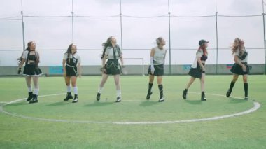 Altı dansçıdan oluşan bir grup çimenli bir alanda koreografili bir gösteri yapıyor. Şık kıyafetler giyiyorlar ve hareketli pozlar veriyorlar. Dans yeteneklerini sergiliyorlar. Arka planda kale direkleri olan bir spor sahası yer alıyor.