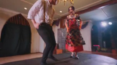 Sahnede geleneksel bir dans sergileyen bir çift. Kadın çiçek desenli renkli bir elbise giyer, erkek ise beyaz gömlek ve siyah pantolon giyer. Arka planda bir avize ve sahne ışıkları var..