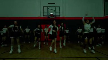 Bir grup dansçı jimnastik salonunda senkronize hareketler sergiliyor. Dinamik ve enerjik bir atmosferi vurgulayarak siyah ve beyaz karışımlı şık kıyafetler giyiyorlar..