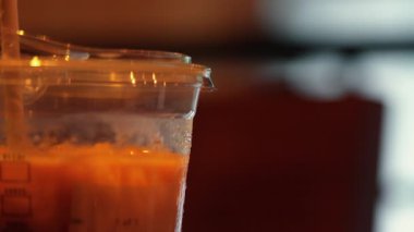 Buzlu kahveyle dolu şeffaf bir fincanın yakın çekimi, yüzeyde görünen bir yoğunlaşma. Arka plan hafifçe bulanık, sıcak ve davetkar bir atmosfer yaratıyor..