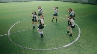 Futbol sahasında dans eden altı genç kadından oluşan bir grup. Aynı kıyafetleri giymişler, enerjik hareketler sergiliyorlar. Arka planda yeşil çim ve kale direkleri görünüyor..