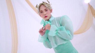 Saf kolları ve papyonu olan, yumuşak, nötr bir zemine karşı zarif poz veren, nane yeşili kıyafetli bir manken. Görünüşü, saçındaki çiçek aksesuarlarıyla tamamlanmış..