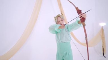 Nane yeşili kıyafetli bir kişi arka planda kumaş örtüsü olan minimalist bir stüdyoda fiyonk çiziyor..