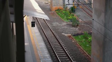 Bir tren istasyonundaki tren yolu manzarası. Kenarlarında otlar ve bitkiler var. Platform kısmen görülebilir ve arka planda bir sinyal görülebilir..