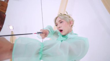 Kabarık kolları ve yayı olan, ok ve yay tutan pastel yeşil kıyafetli bir adam. Hassas bir tacı var ve hafif ışıklandırmayla tuhaf bir ortamda..