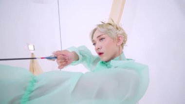 Saf kumaşlı, açık yeşil kıyafetli, ok ve yay tutan, minimalist beyaz bir arka plana ayarlanmış biri. Bireyin kısa sarı saçları ve çiçekli bir başlığı var. Sakin ama odaklanmış bir ifade yayıyor..