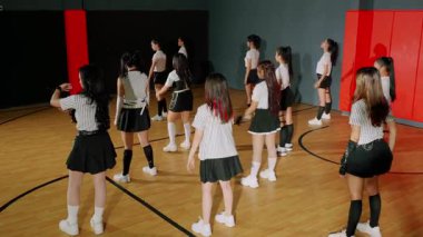 Bir spor salonunda eşleşen bir grup dansçı antrenman yapıyor. Siyah etekler ve çizgili bluzlar giyiyorlar ve koreografili bir gösteri sergiliyorlar. Kurulumda ahşap zemin ve renkli duvarlar yer alıyor..