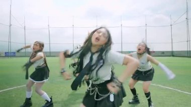 Bir futbol sahasında senkronize dans yapan altı kızdan oluşan bir grup. Spor üniformaları giyiyorlar ve dairesel bir şekilde konumlanmışlar. Arka planda açık bir gökyüzü ve bir futbol hedefi yer alıyor..