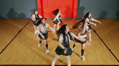 Bir spor salonunda dans gösterisi yapan altı genç kadından oluşan bir grup. Etekleri ve spor ayakkabıları ile uyumlu kıyafetler giyiyorlar, enerjik hareketler sergiliyorlar. Arka planda kırmızı bir duvar ve ahşap bir zemin var..