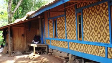 Bambu ve ahşaptan yapılmış geleneksel bir ev. Duvarlarda karmaşık dokuma desenleri var. Ev verandalı ve ahşap kapılı yeşilliklerle çevrili..