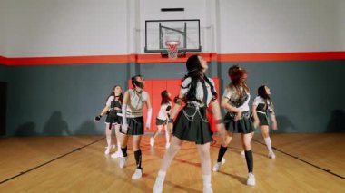 Bir grup dansçı jimnastik salonunda senkronize hareketler sergiliyor. Etek ve spor ayakkabılarla uyumlu kıyafetler giyiyorlar. Arka planda basketbol potası var..
