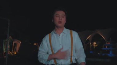 Beyaz gömlek ve omuz çantası giymiş, şaşırmış bir ifadeye sahip genç bir adam, karanlık bir ortamda duruyor. Arka plan bulanık, hareket ya da aciliyet belirtisi var..