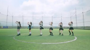 Bir futbol sahasında dans eden altı dansçıdan oluşan bir grup enerjik koreografi sergiliyor. Şık kıyafetler giyiyorlar ve arkalarında bulutlu bir gökyüzü olan dairesel bir düzende konumlanmışlar..