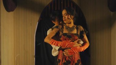 Klasik bir ortamda bir erkek ve bir kadının olduğu dramatik bir sahne. Kadın siyah süslemeler ve turuncu eldivenlerle zarif bir kırmızı elbise giyer, erkek ise beyaz gömlek giyer. Işık dengesiz bir atmosfer yaratıyor.