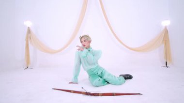 Nane yeşili elbiseli bir kişi beyaz bir zeminde zarif bir şekilde poz verir, önünde bir fiyonk yatar. Arka planda yumuşak perdeler ve ışıklar var, sakin bir atmosfer yaratıyor..