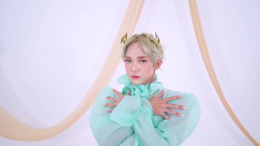 Nane yeşili kıyafetli, geniş fiyonklu ve kabarık kollu, hafif bir arka planda akıcı kumaşla zarif poz veren biri..