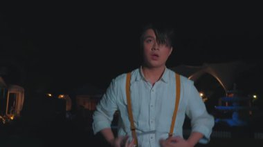 Koyu renk saçlı ve pantolon askısı olan genç bir adam gece dikkatlice yukarı bakar. Arka planda hafif ışıklar ve loş bir açık hava ortamı ile çevrili..