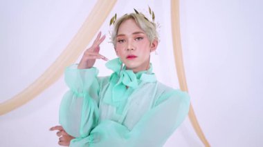 Kafasında çiçek aksesuarlarıyla süslenmiş, akıcı tasarımı olan yeşil bir elbise giyen bir adam. Arka planda yumuşak perdeler var, ruhani bir atmosfer yaratıyor..