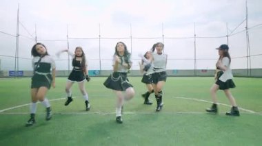 Bir spor sahasında senkronize dans yapan altı kızdan oluşan bir grup. Birbirlerine uygun kıyafetler giymişler. Bulutlu bir gökyüzünün arka planına karşı koreografilerini sergiliyorlar..