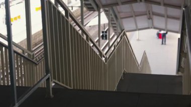 Modern bir tasarım ve parmaklıklarla bir tren istasyonuna giden merdiven manzarası. Sahne çoğunlukla boş, platformda yürüyen bir kişi var..