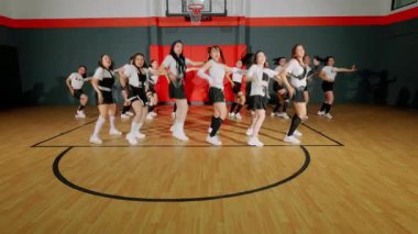 Bir grup dansçı bir basketbol salonunda koreografi gösterisi yapıyor. Gösterişli kıyafetler giyiyorlar ve enerjik hareketler sergiliyorlar. Ön planda dansçılar ve arka planda diğerleri var..