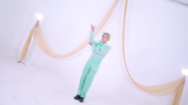 Nane yeşili kıyafetli bir insan minimalist bir stüdyoda yumuşak perdelerle poz verir. Arka plan beyaz, temiz ve modern bir estetik yaratıyor..