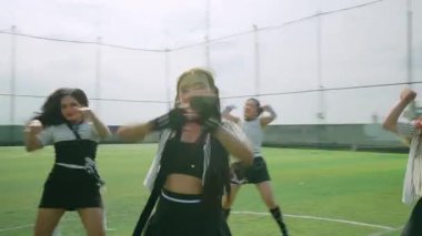 Bir grup dansçı açık hava sahasında, çizgili gömlek ve eteklerle şık kıyafetler giyerek gösteri yapıyorlar. Sahne dinamik hareketleri ve ifadeleri yakalıyor, enerji ve takım çalışması sergiliyor.