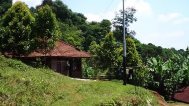 Yeşillik ve ağaçlarla çevrili geleneksel bir evin bulunduğu sakin bir kırsal alan. Gökyüzü birkaç bulutla parlak mavi, barışçıl bir atmosfer yaratıyor..