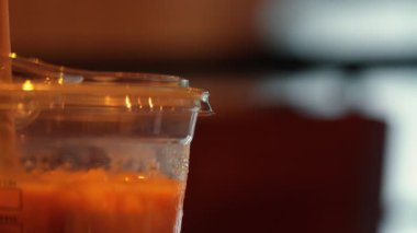 Buzlu içecekle dolu şeffaf bir fincanın yakın çekimi, dışarıdan görülebilen bir yoğunlaşma. Arka plan hafifçe bulanık, sıcak bir ortam yaratıyor..