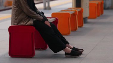 Tren istasyonunda kırmızı bir taburede oturan ve arka planda birden fazla kırmızı tabure olan biri. Sahne, bireyin ayaklarına ve günlük kıyafetlerine odaklanmış bir bekleme anını yakalıyor..