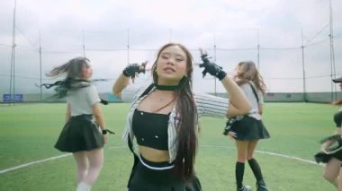 Altı dansçıdan oluşan bir grup yeşil bir çim sahasında koreografili bir gösteri yapıyor. Şık kıyafetler giyiyorlar, etek ve spor kıyafetleri sergiliyorlar. Arka planda bulutlu bir gökyüzü ve bir spor ağı yer alıyor..