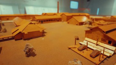 Tarihi bir bina kompleksinin detaylı mimari modeli, evler, depolar ve ağaçlar gibi çeşitli yapılar içeriyor, hepsi de ahşaptan yapılmış ve kumlu bir zeminde sergileniyor..
