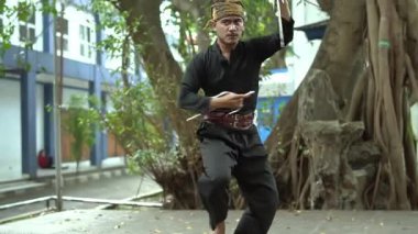 Geleneksel kıyafetli bir sanatçı, ağaçlar ve şehir mimarisi ile çevrili açık havada bir kılıçla dövüş sanatları dansını gösterir..