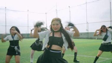 Bir grup dansçı bir spor sahasında, enerjik koreografi sergiliyorlar. Siyah bir bluz ve çizgili bir ceket giyen baş dansçı anlamlı hareketlerle göze çarpıyor. Arka planda parlak bir gökyüzü ve bir futbol sahası var..