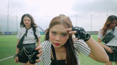 Tarz sahibi kıyafetler giymiş üç genç kadın arka planda bulutlu bir gökyüzü olan bir futbol sahasında duruyor. Merkezdeki figür uzun saçlı ve kendinden emin bir ifadeye sahipken, diğerleri onun yanında poz veriyor..