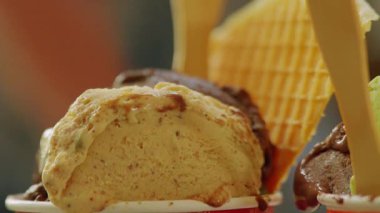 Renkli bardaklardaki dondurma kepçelerinin yakın çekimi, kremsi bir dokuya ve çeşitli tatlara sahip. Bir kepçe fındıklı açık kahverengi, diğeri koyu çikolatalı. Waffle konileri arka planda görünür..