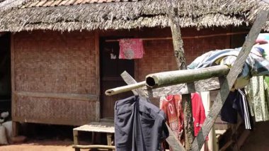 Ahşap bir kapı ve verandası olan sazdan çatılı bir bambu evi. Giysiler yakınlarda bir hatta asılı, basit bir kırsal yaşam tarzını sergiliyorlar..
