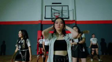 Bir grup dansçı, enerjik koreografi sergileyerek bir spor salonunda gösteri yapıyorlar. Siyah bir bluz ve çizgili bir gömlek giyen baş dansçı ön planda, diğerleri ise arka planda şık dans kıyafetleri içinde..