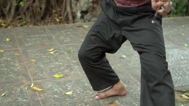 Siyah gömlek giyen ve eşsiz bir kestane rengi kemer takan biri açık havada dinamik bir hareket sergiliyor. Arka planda yeşillik var, doğal bir ortam var..