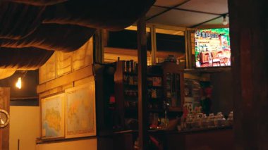 İçinde ahşap mobilyalar, duvarlarda haritalar ve renkli bir sahne gösteren bir televizyon bulunan bir kafe ya da barın içi. Ortam yumuşak ışıklandırmayla sıcak..