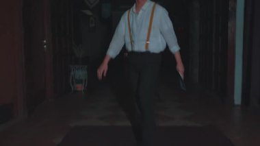 Pantolon askısı olan bir adam loş bir odada yürüyor, muhtemelen tarihi ya da temalı bir ortamda. Arka planda dekorasyonlar ile atmosfer gizemli..