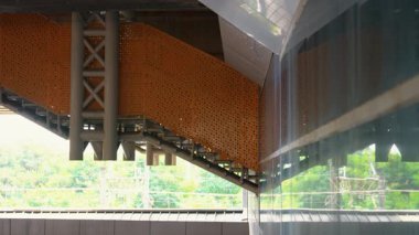 Modern bir mimari detay turuncu delikli panelleri olan bir merdiven sergiliyor, cam yüzeyleri yansıtıyor ve arka planda yeşillik var..